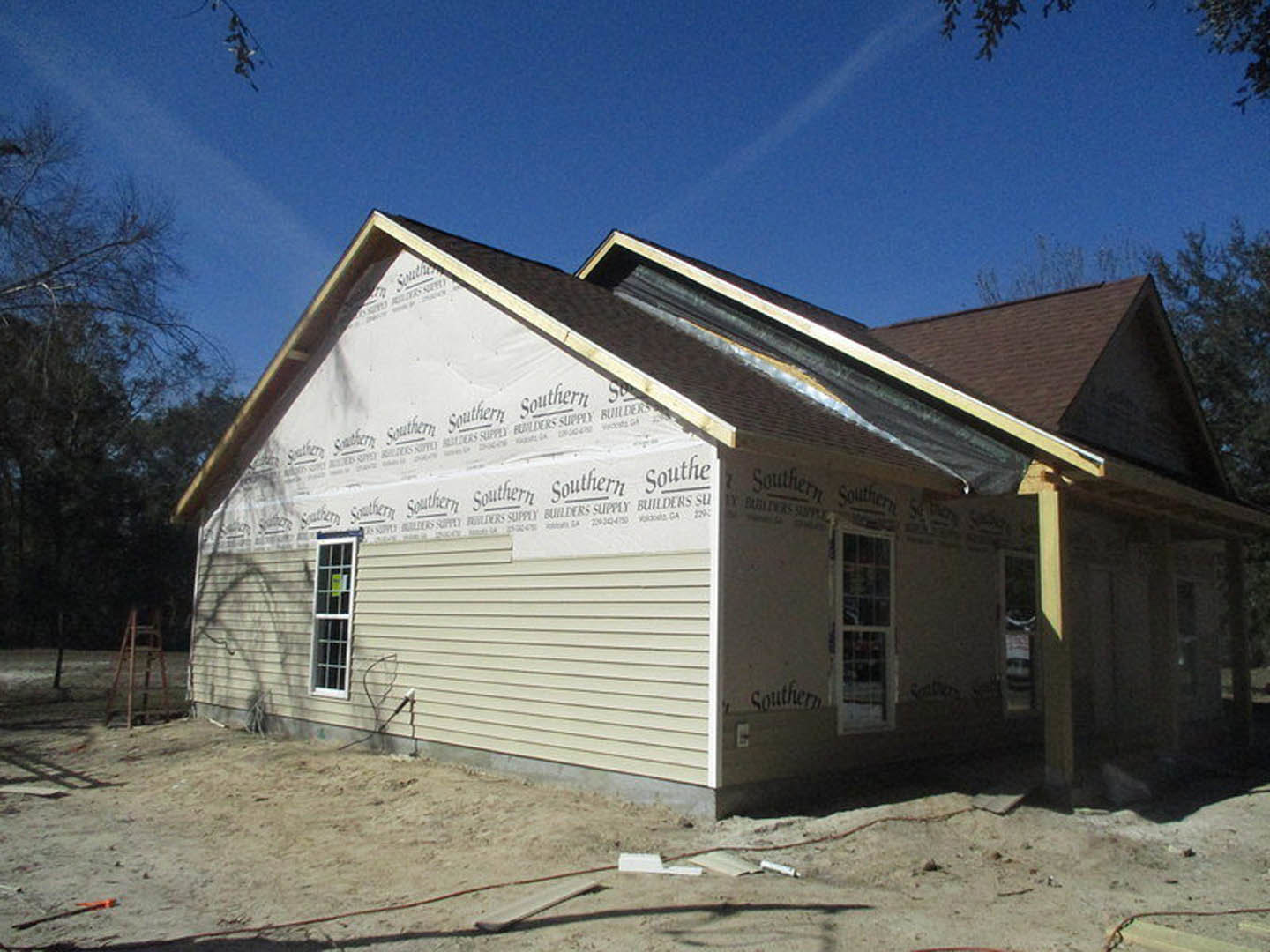 Framed house under construction with installed roof and insulation, window displaying a sign, leafless tree nearby, blue sky with scattered clouds, pile of lumber on dirt ground