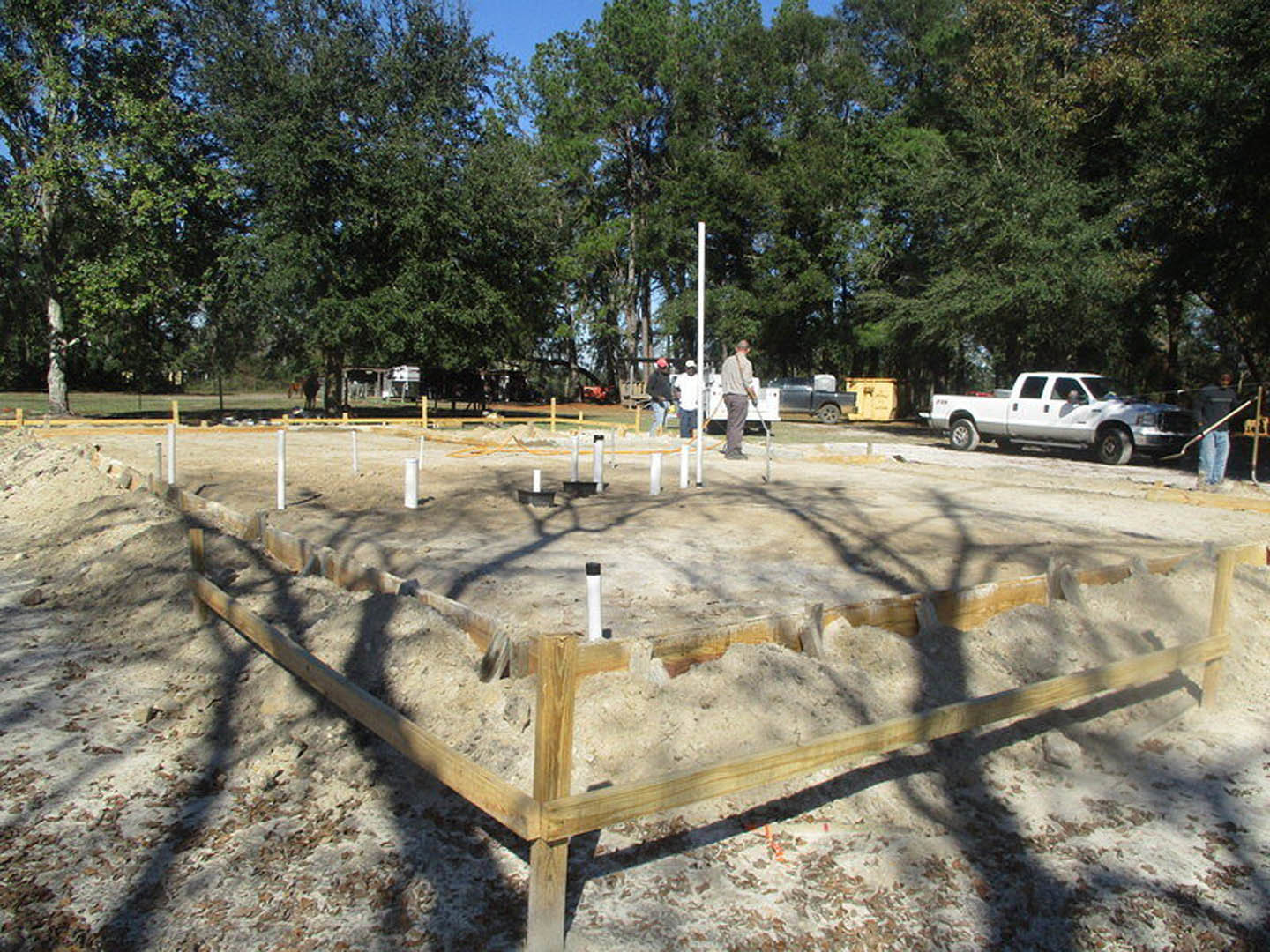 Snow-covered construction site with scattered wooden planks, pile of sand, white pickup truck, and several people working outdoors near trees under a cloudy sky