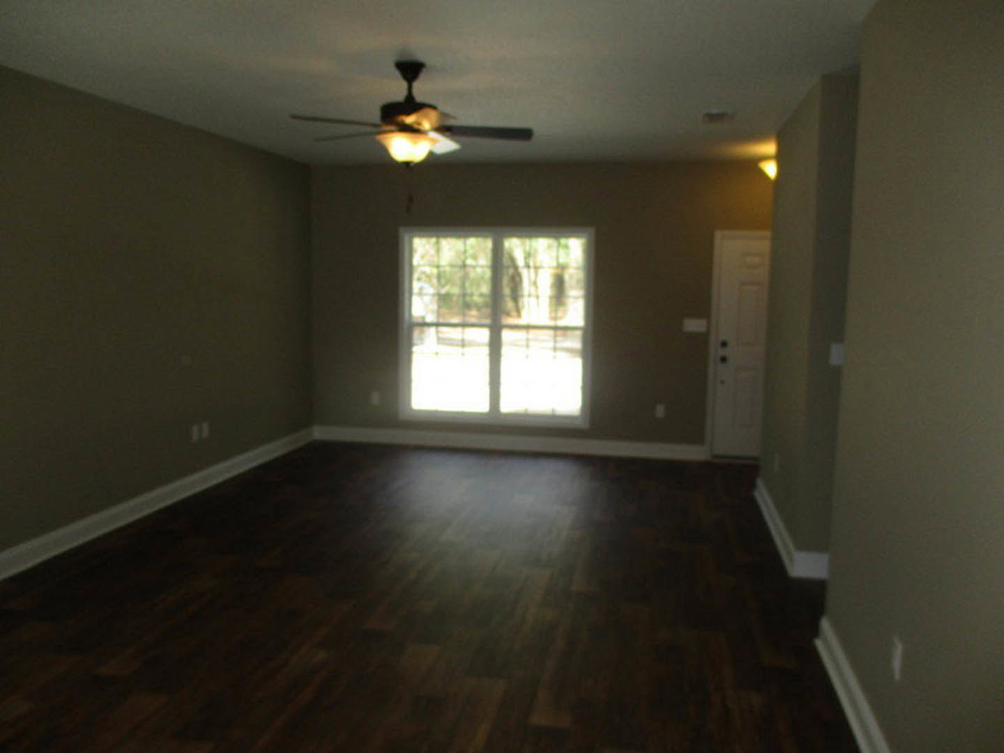 Dark wood flooring, white walls, ceiling fan with light fixture, large window showing outdoor greenery, partially visible white door with black hardware.