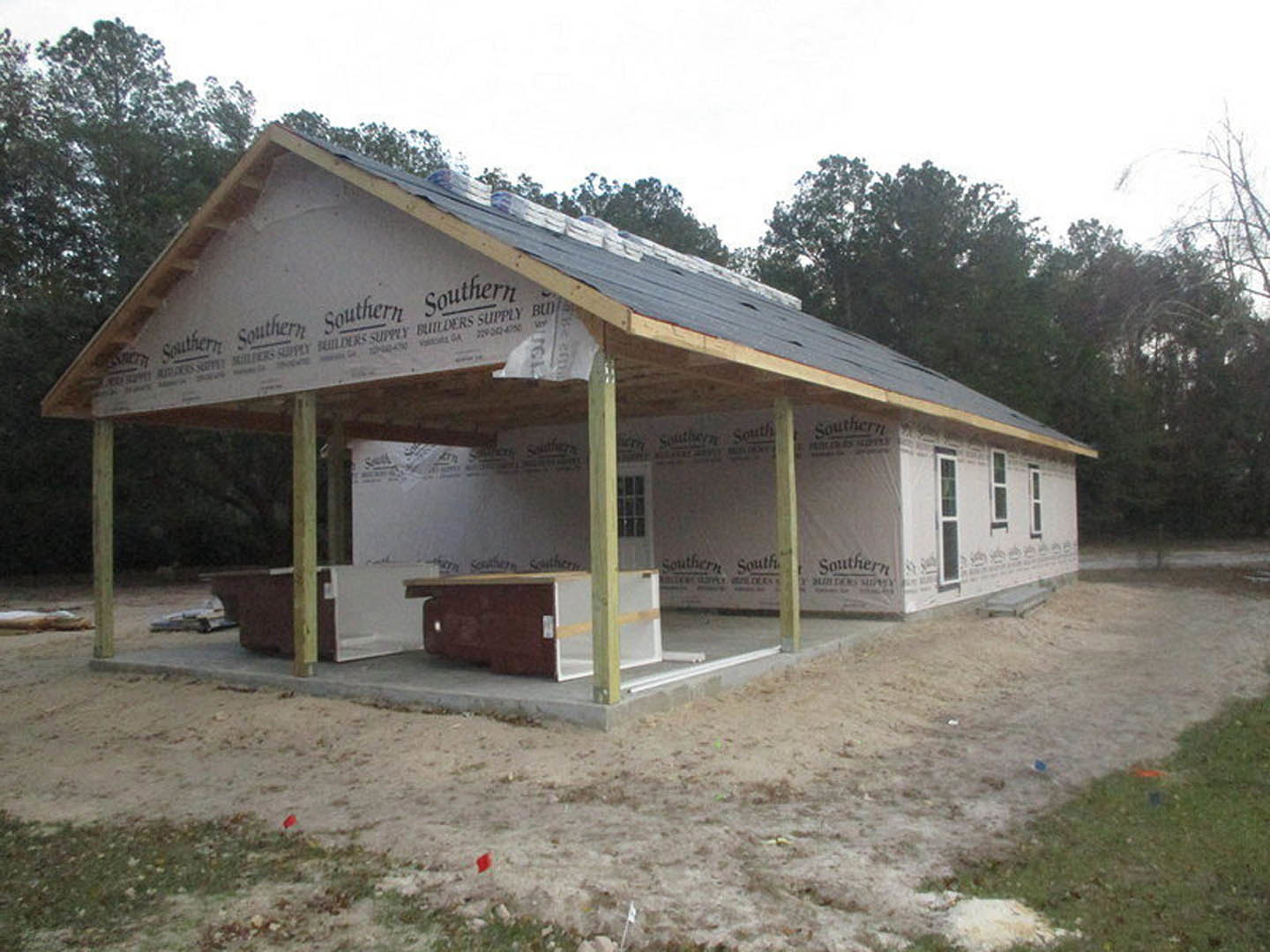 Wood-framed house under construction with covered porch, exposed beams, unfinished roof, and construction materials scattered on site