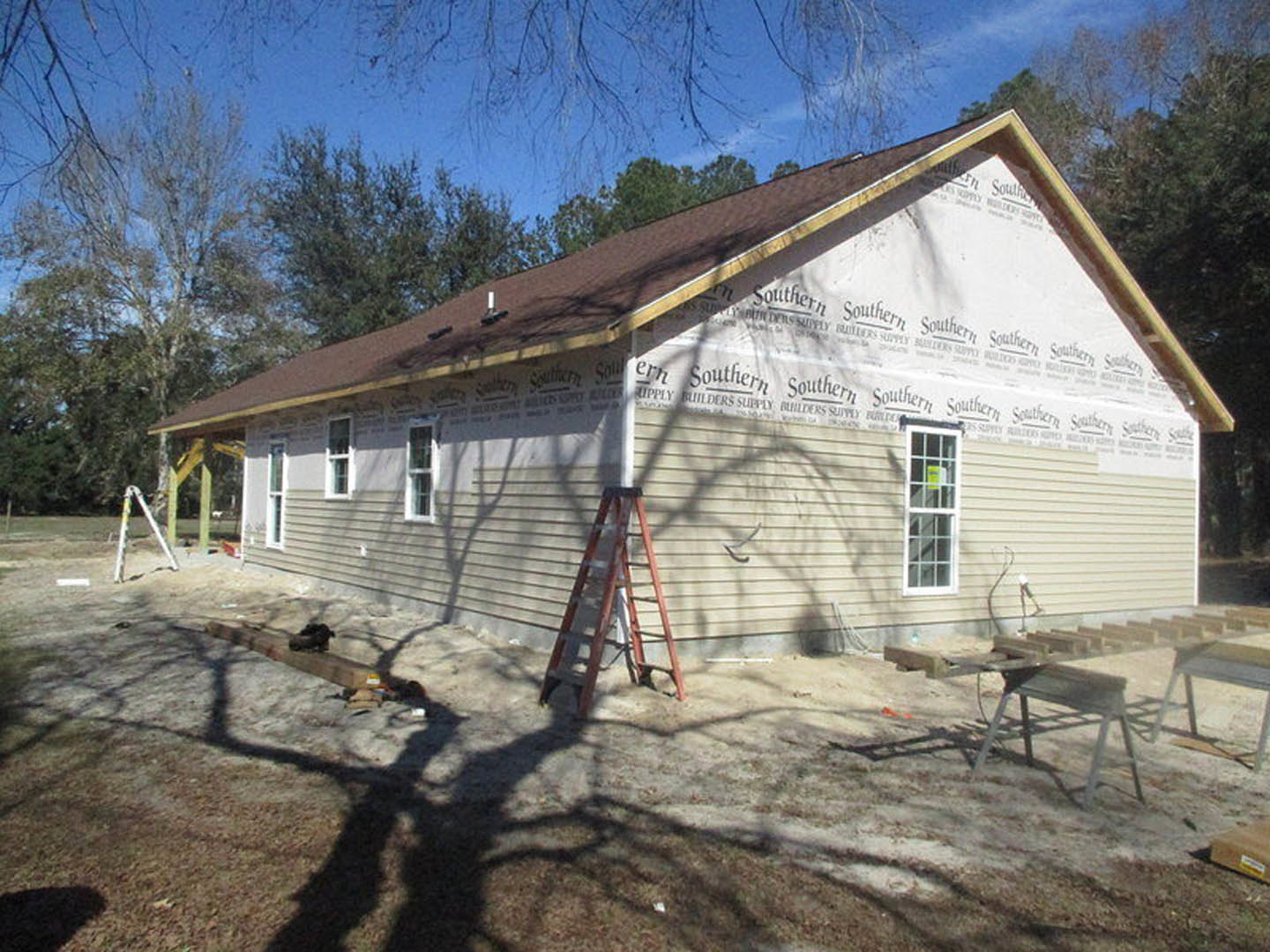 Partially constructed house with exposed siding, white ladder leaning against wall, dirt yard, window displaying sign, metal bench nearby, trees in background
