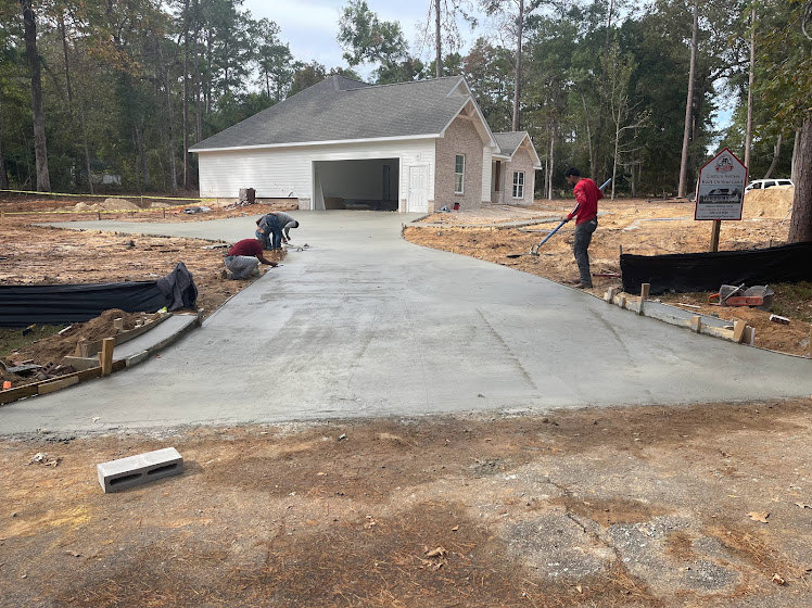 Workers installing or repairing concrete driveway in front of modern house with white siding, surrounded by green lawn, trees, and landscaping under blue sky