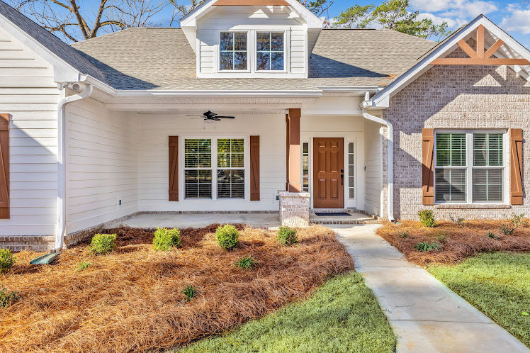 Two-story home with light siding, covered front porch, large windows, manicured lawn, concrete walkway, and blue sky