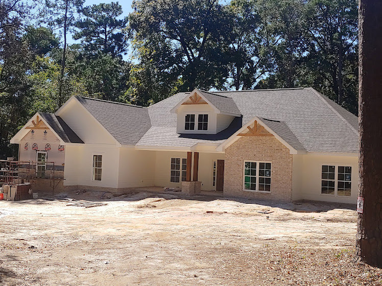 Wood-framed house under construction surrounded by tall trees, partially finished roof, exposed walls, and open window spaces