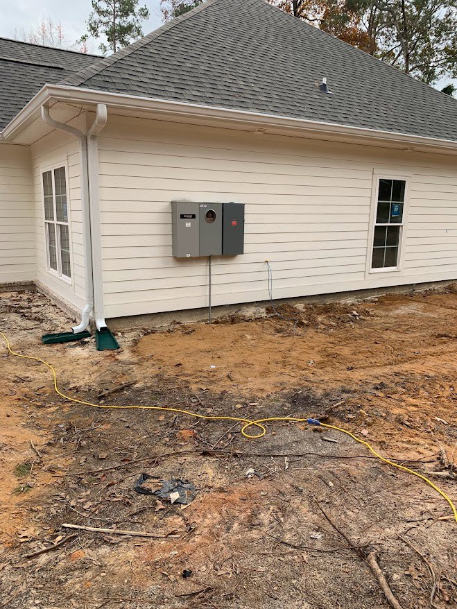 Light-colored siding house with white-framed window, grey utility boxes mounted on exterior wall, yellow hose lying on dirt near foundation, small tree and shed visible in