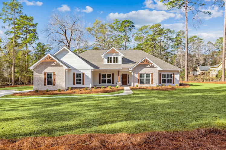 Two-story home with light siding, covered front porch, manicured green lawn, mature trees in the background, and partly cloudy sky overhead