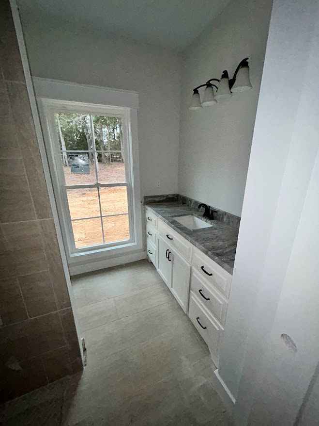 Bathroom featuring a rectangular window above a white sink with stone countertop, tile backsplash, wood cabinetry, and light-colored floor tiles