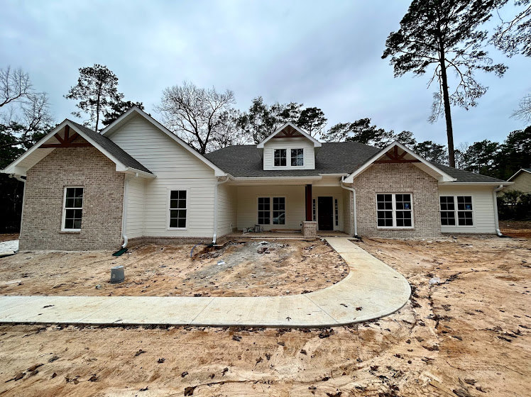 Two-story house under construction with exposed framing, white window frames, dirt driveway, leafless tree, and concrete sidewalk surrounded by bare ground and trees in the