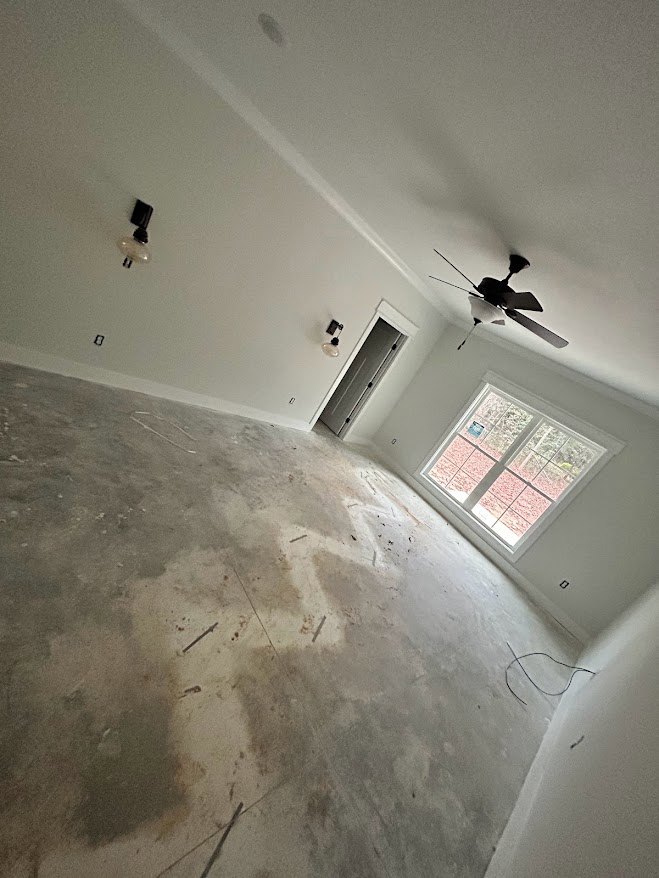 Neutral-toned room with plaster walls, ceiling fan with light fixture, large window allowing daylight, and door with wall-mounted light.