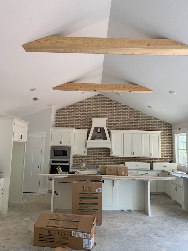 White kitchen cabinets with silver handles, exposed wooden ceiling beams, white oven with illuminated center, light countertops, stainless steel sink, and cardboard box with