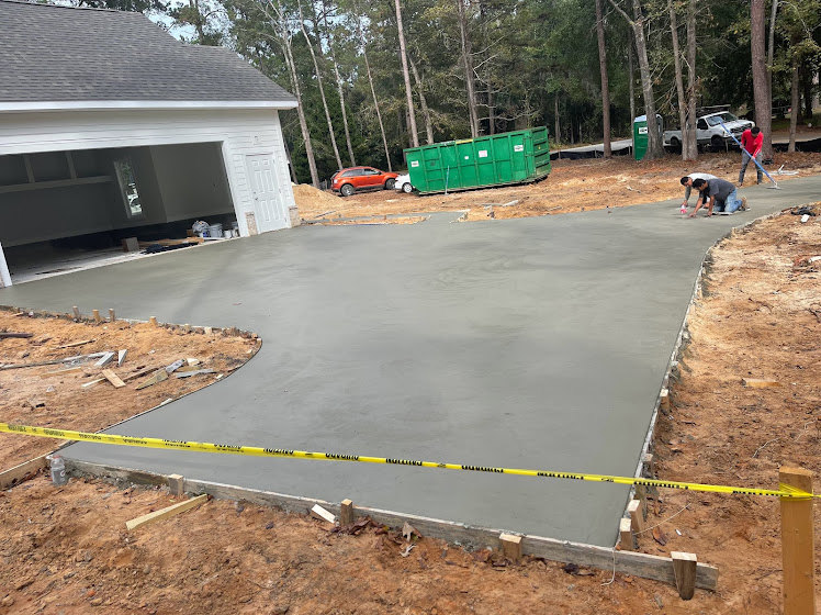Man standing on unfinished concrete slab foundation beside green dumpster with white labels; surrounding area includes asphalt road, trees, and construction materials.