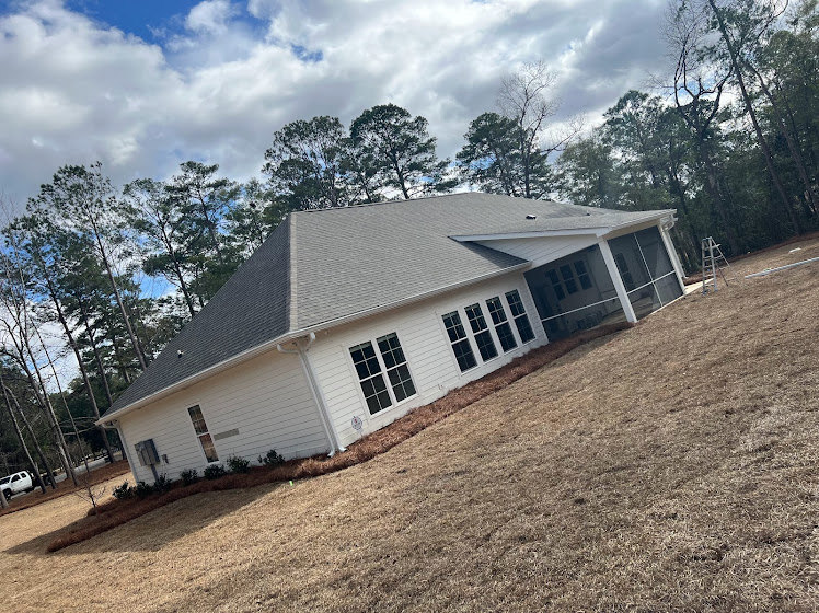 Two-story house with white siding and dark roof, large windows, surrounded by expansive green lawn and mature trees under partly cloudy sky