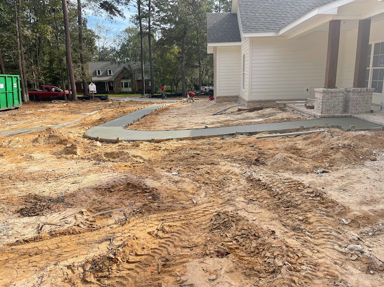Modern home with light siding, paved driveway, exposed soil around foundation, mature trees in background, clear blue sky