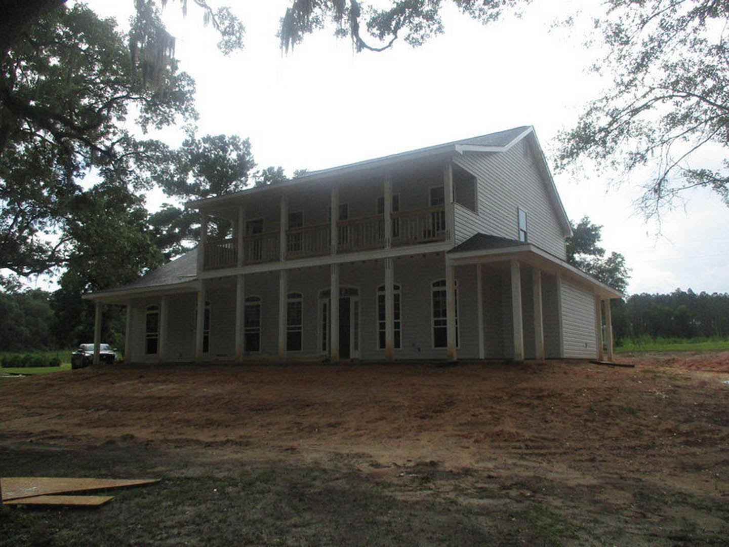 Spacious white house with covered porch, unfinished dirt yard, multiple windows, and surrounding trees; construction materials visible near foundation