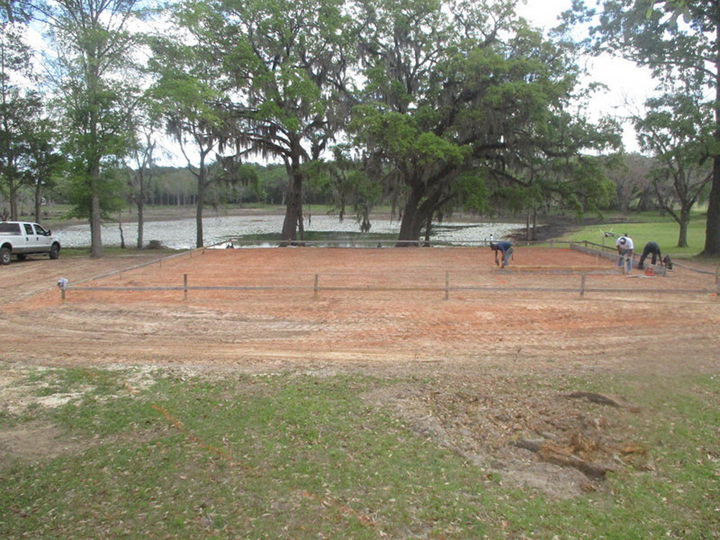 Tennis court surrounded by grass and trees, two people playing tennis, white truck parked nearby, moss-covered trees in background