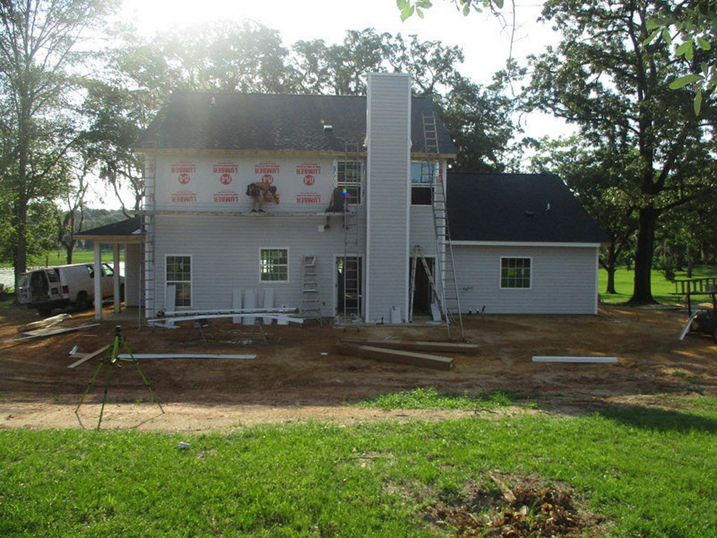 Wood-framed house under construction with exposed sheathing, a man working on a ladder near multiple square windows, grassy yard, white van parked with door open, cow standing