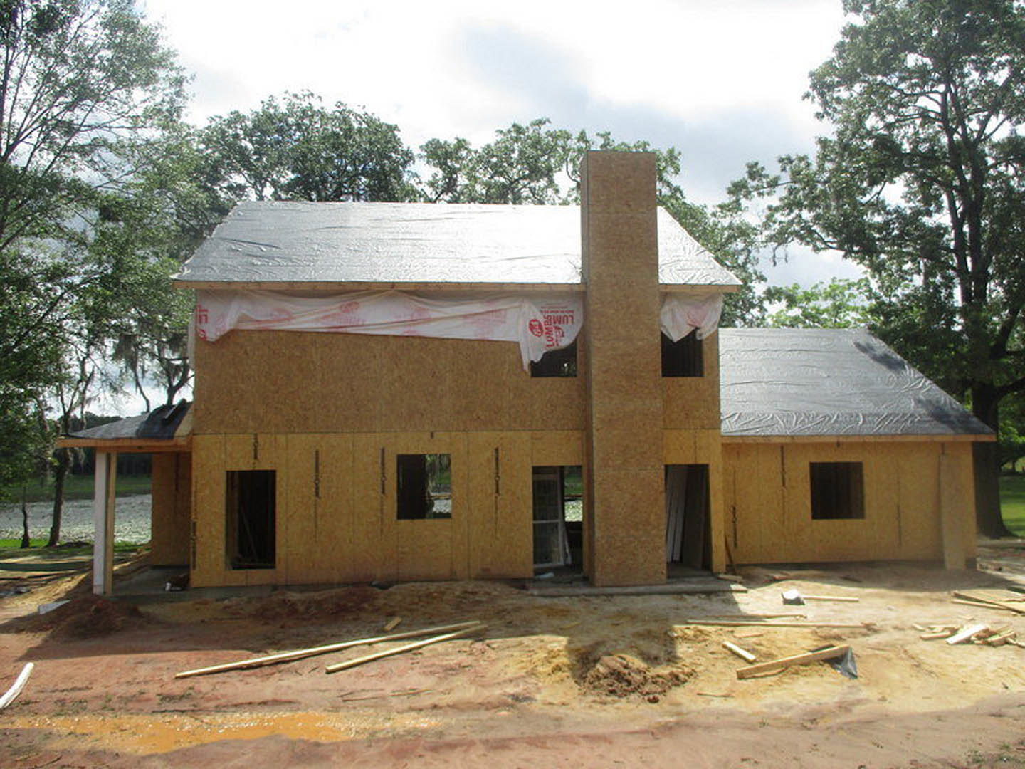 Framed house under construction with exposed plywood walls, chimney, square-paned window, surrounded by trees and blue sky