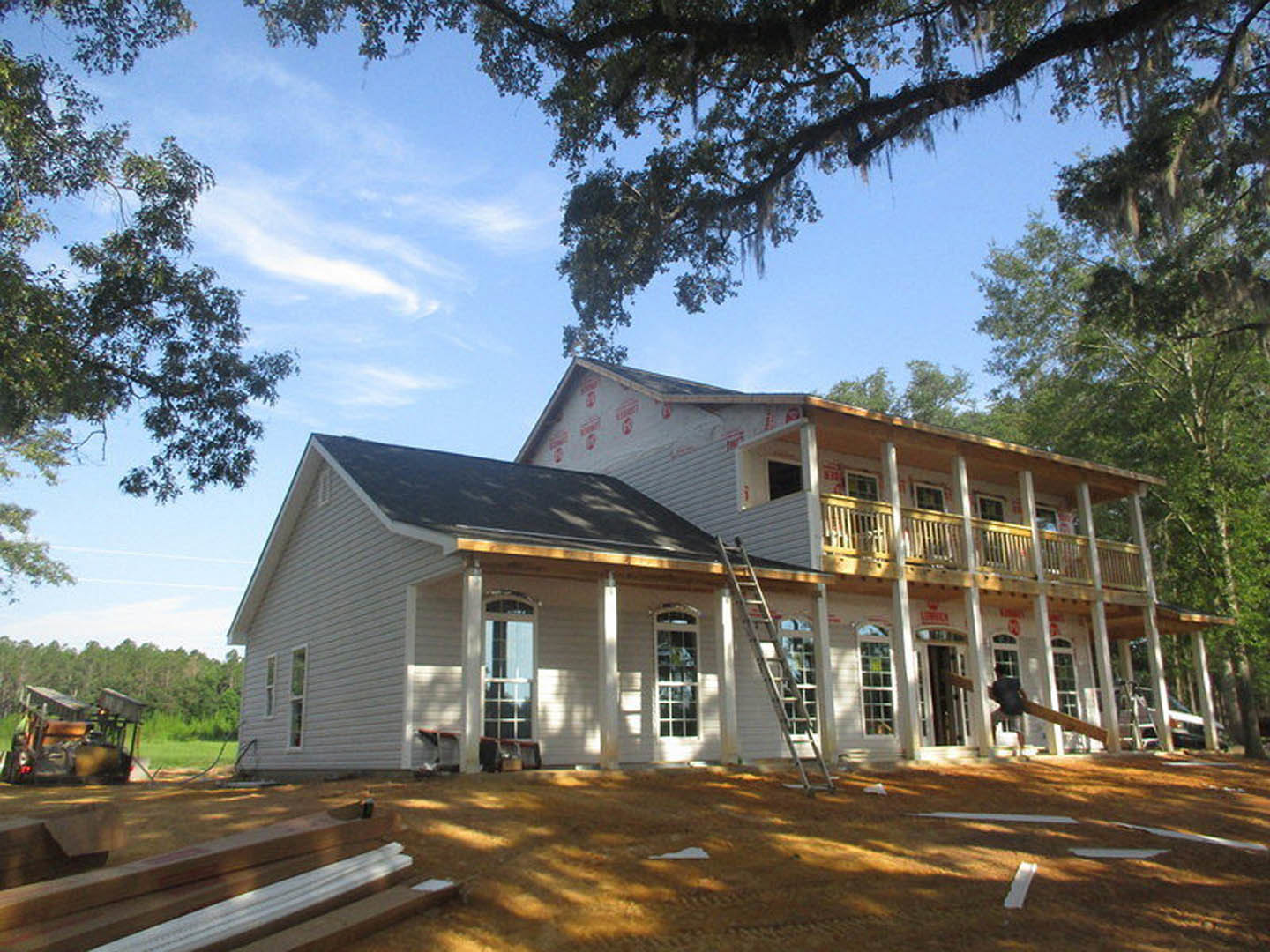 Wood-framed house under construction with multi-pane windows, white trim, exposed beams, ladder leaning against siding, large leafy tree in foreground, cloudy sky overhead, truck
