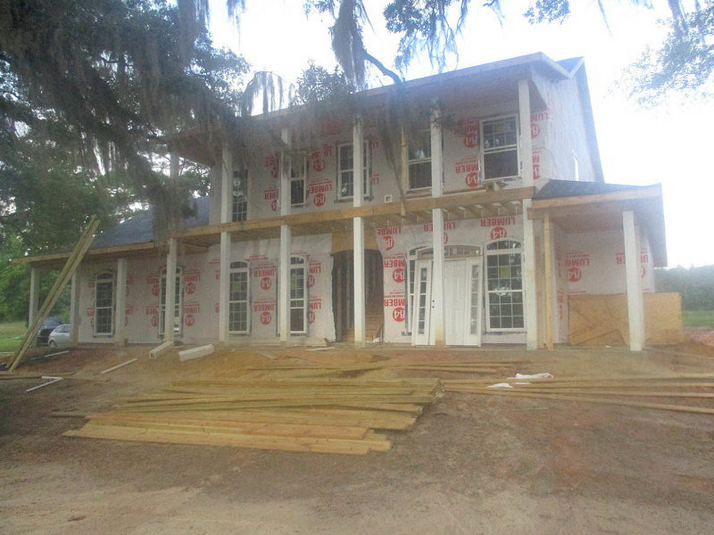 Framed wooden house under construction with exposed beams, unfinished porch, parked cars in front, surrounding trees, and scattered lumber on the ground