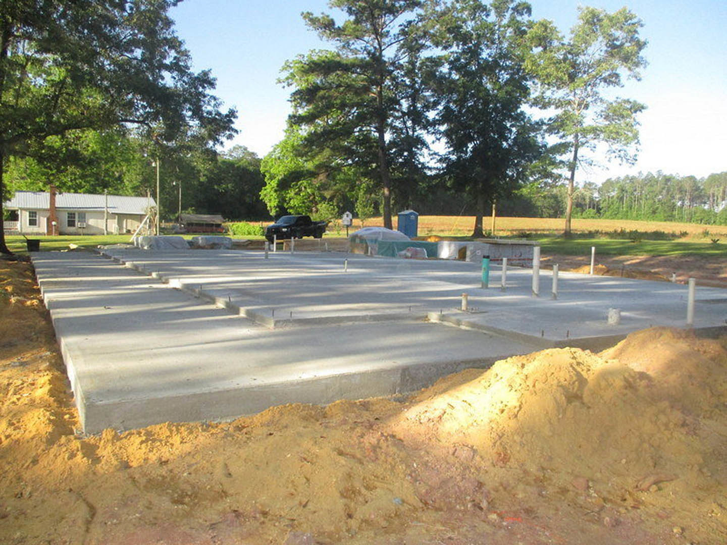Partially built house with exposed framing, dirt lot, black van parked on asphalt road, surrounding trees, and visible chimney and window.