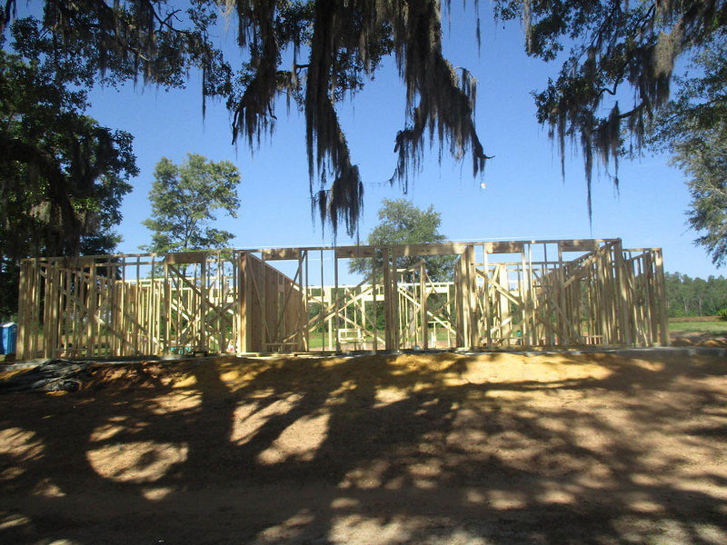 Wooden house frame under construction with tree casting shadows on dirt ground, moss hanging from branches, partially visible window, and surrounding greenery.