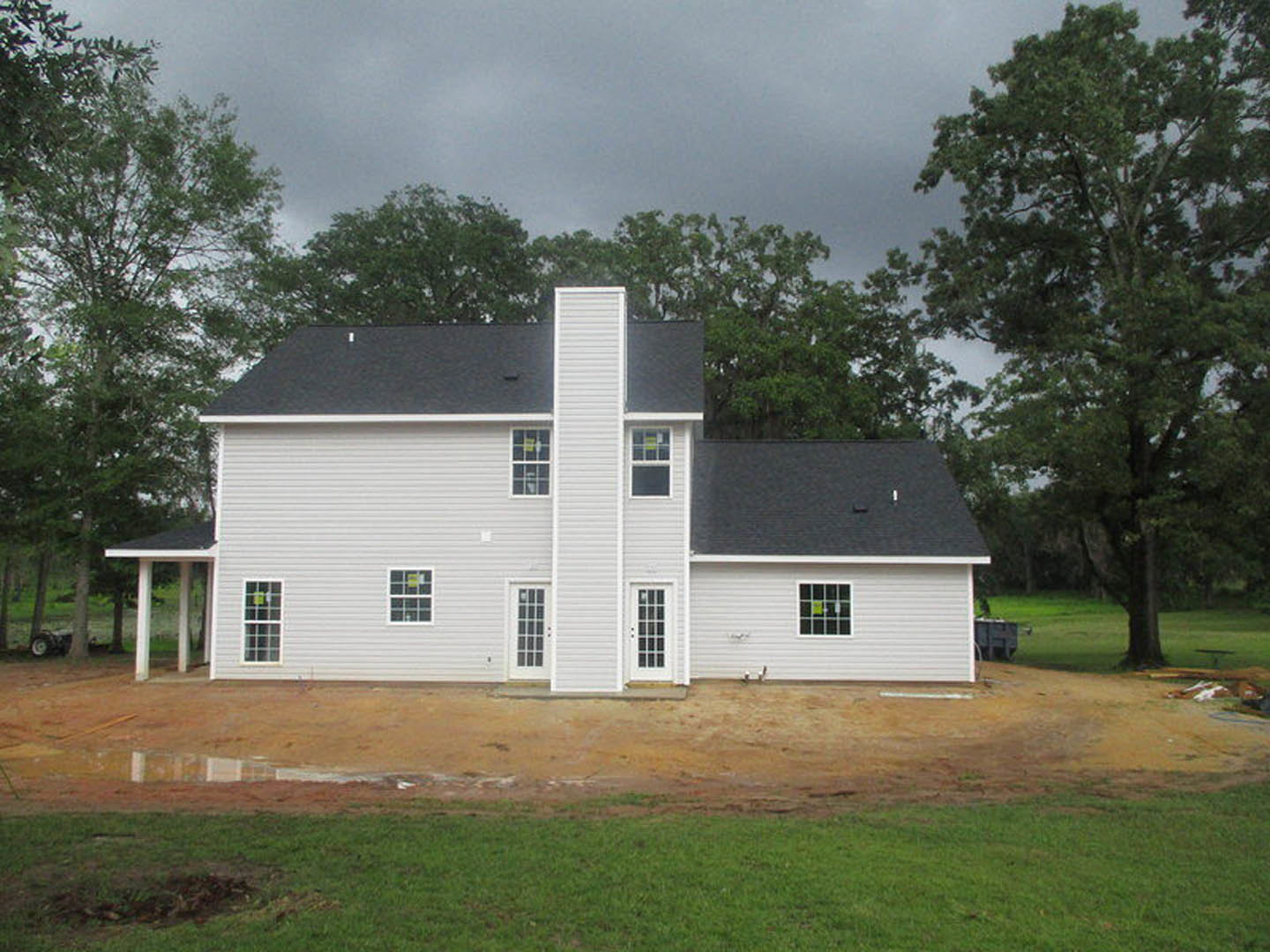 White siding house under construction with chimney, multiple windows, and white door, surrounded by green trees and grassy lot.