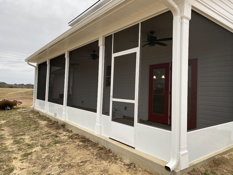 White house with horizontal siding, red front door with glass window, covered porch featuring exposed wooden beams, ceiling fan with light, multiple windows, green lawn in