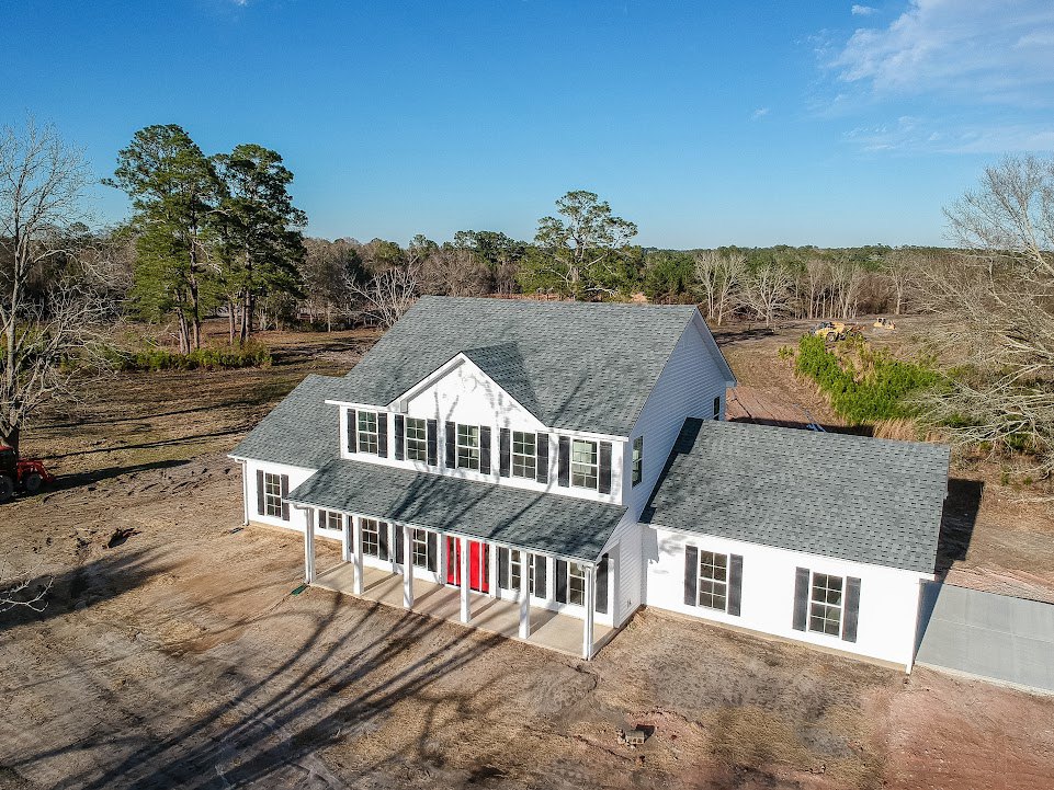 White two-story house with red front door, black shutters, multiple windows, manicured lawn, mature trees, and partly cloudy blue sky