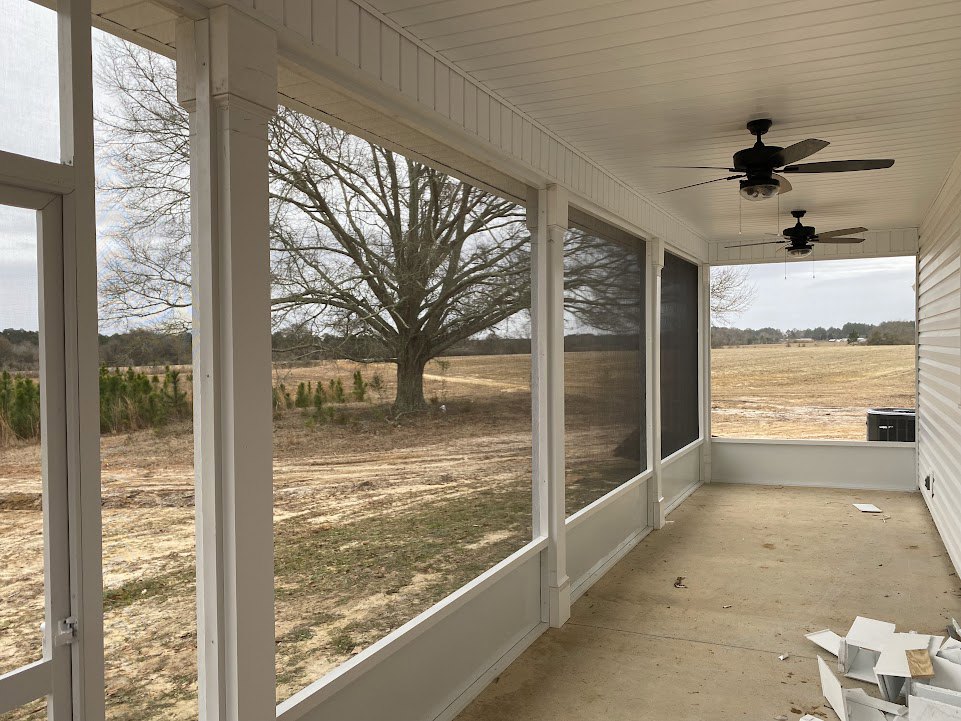 Covered porch with concrete floor, ceiling fan with light, large window overlooking grassy yard, tree, and fence; pile of broken white boxes near building exterior.