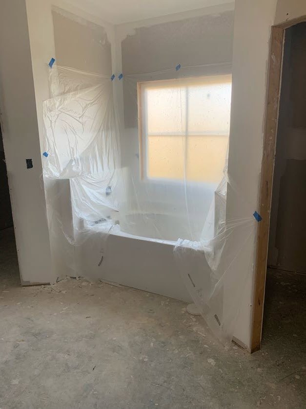 Bathroom under construction with plastic sheeting covering walls, exposed concrete floor scattered with debris, window, unfinished tub, and door partially wrapped in plastic.