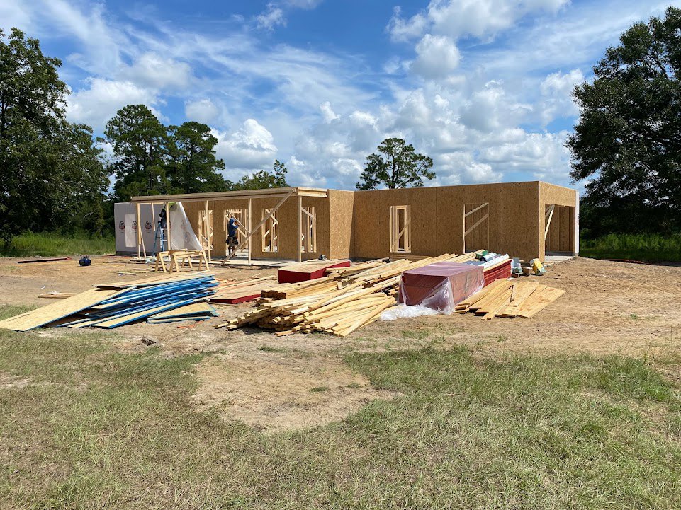 Framed wooden house under construction with exposed beams, stacks of lumber, plastic-wrapped materials, person standing inside, grassy yard, trees, and partly cloudy blue sky