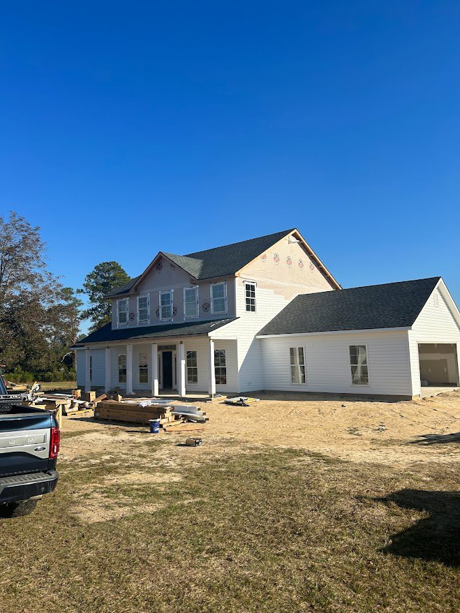 Partially built house with exposed framing, roof shingles, construction materials and pallets scattered in the yard, pickup truck parked nearby, mature trees and blue sky in the