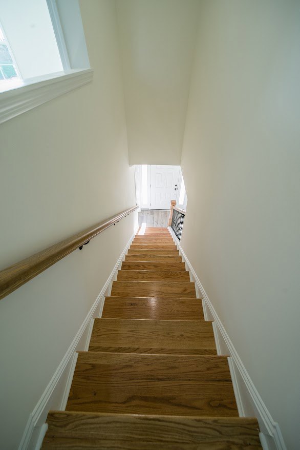 Wooden staircase with matching handrail ascending beside a smooth white plaster wall, natural wood flooring and ceiling visible, window light illuminating the space.