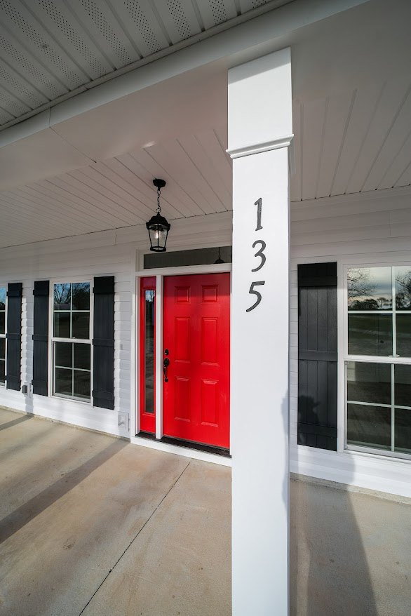Red front door with white column and white trim, adjacent window with white frame, concrete entry floor