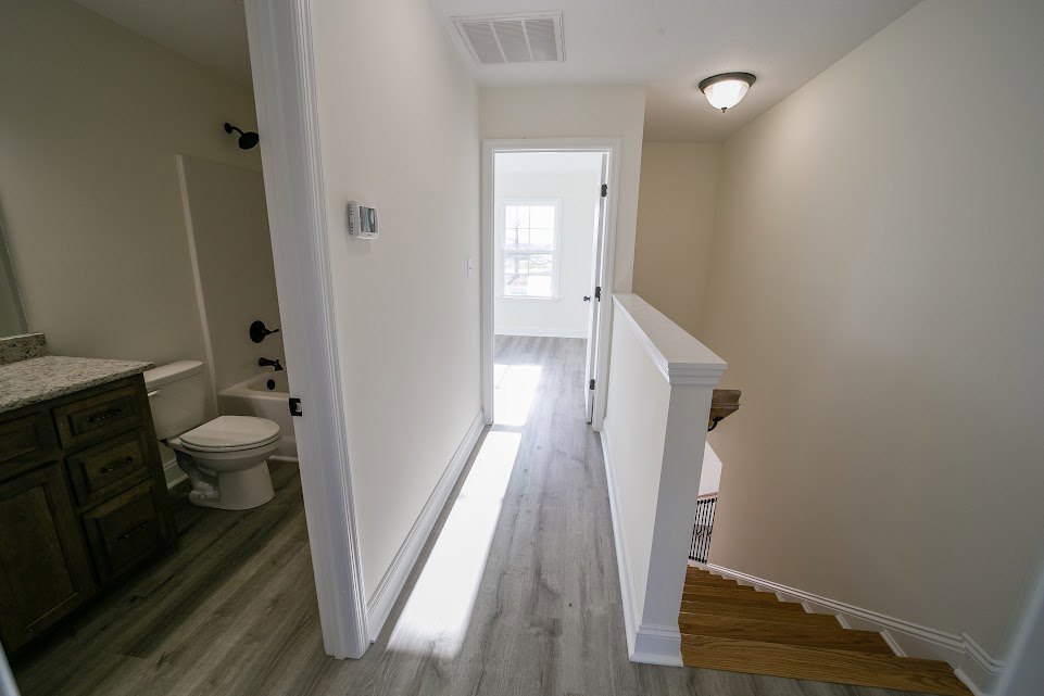 Hallway with light tile flooring, white plaster ceiling and trim, visible staircase, bathroom with toilet and tub, wooden drawer, and partial view of a bed