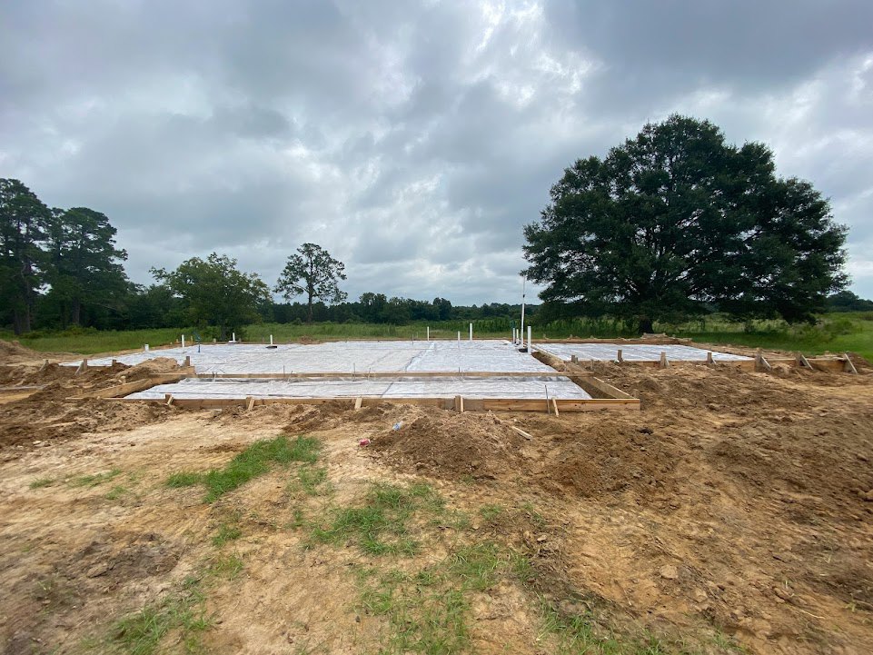 Concrete house foundation surrounded by dirt and tarps, large tree in grassy field, cloudy sky overhead, construction workers on site