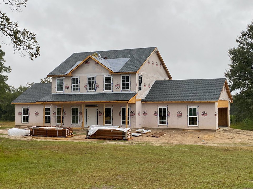 Framed house under construction with exposed wood, grassy yard scattered with pallets, tree with green leaves, overcast sky