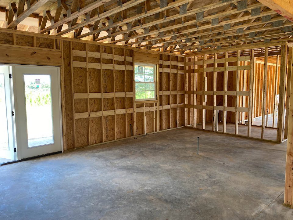 Open room with exposed wood ceiling beams, concrete floor, white-framed window letting in natural light, and a door featuring a window and small sign.