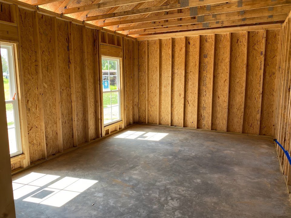 Concrete floor with square tile pattern, exposed wooden ceiling beams, large window letting in natural light, white walls, and visible insulation.