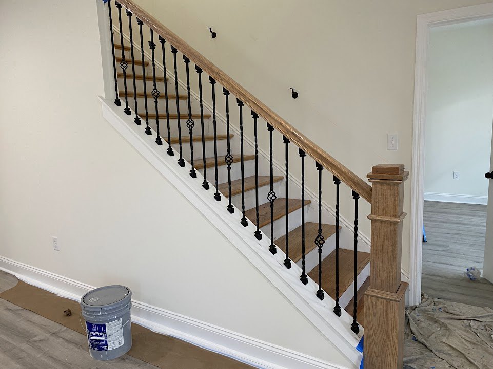 Staircase with black metal railings, wooden post, paint bucket and dirty cloth on unfinished floor in residential interior
