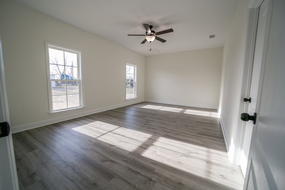 Sunlit room with laminate wood flooring, white plaster walls, ceiling fan with light fixture, multiple windows, and a close-up of a brushed metal door knob