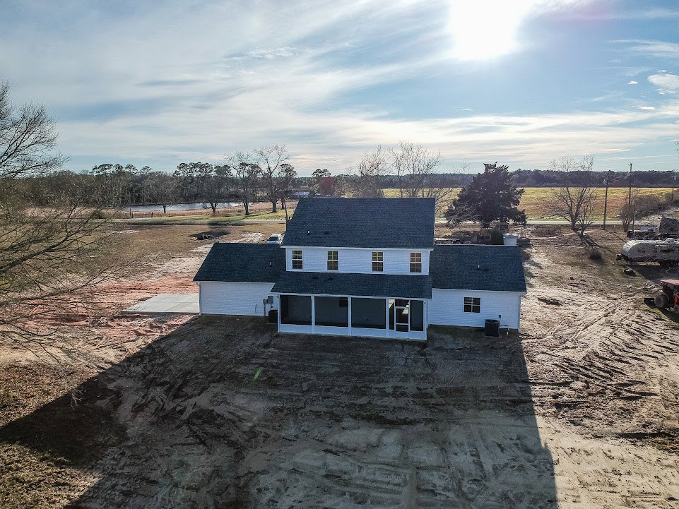 Framed house under construction with covered porch, garage door, dirt driveway, surrounded by grassy field and scattered trees under blue sky with clouds
