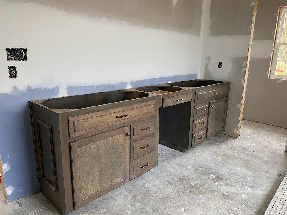 Light wood cabinetry with multiple drawers, stone countertop, stainless steel sink, large window with white trim, neutral walls, and hardwood flooring in a modern kitchen.