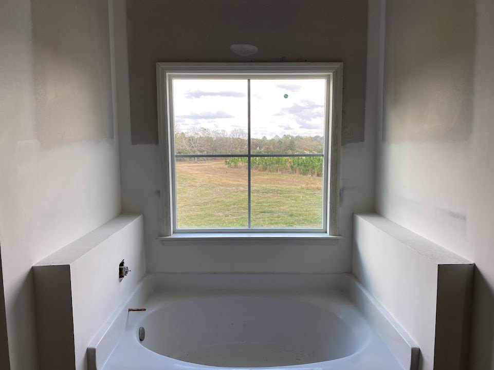 Freestanding white bathtub beneath large window overlooking field and trees, white walls, chrome faucet, adjacent sink and mirror