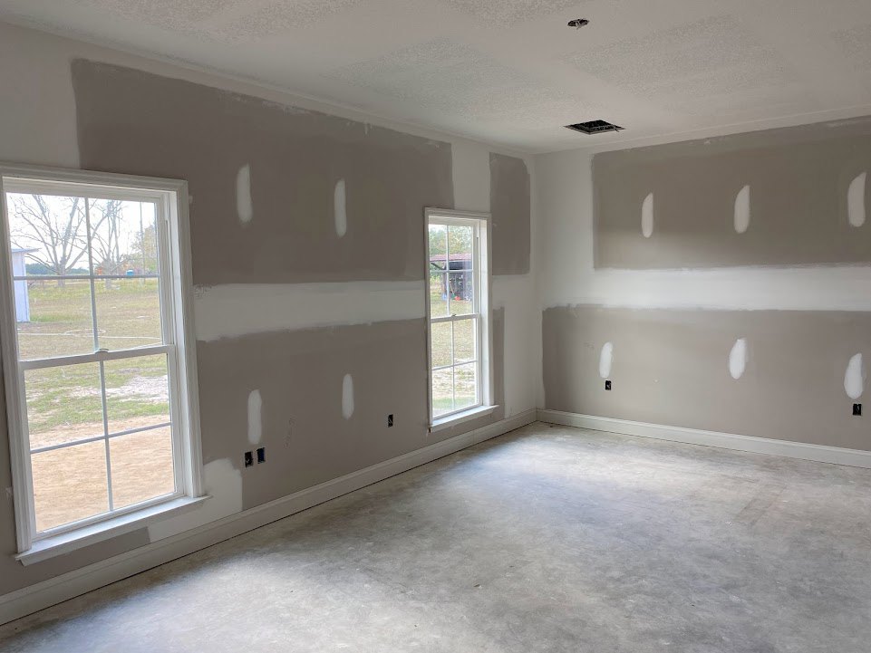 Bright room with smooth white walls, large white-framed window overlooking green grass and trees, light-colored flooring, and ceiling with recessed fixtures