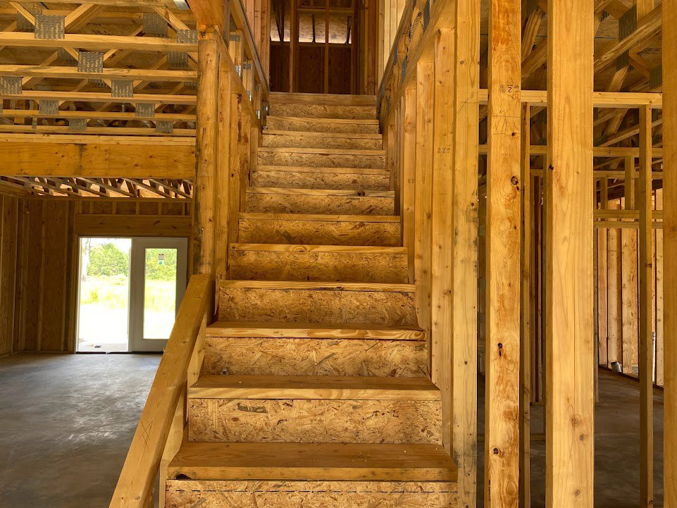 Wooden staircase with matching wood frame, glass-paneled door, large window showing trees and greenery outside, light-colored plank flooring, exposed wood beams on ceiling and