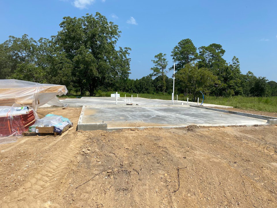 Framed custom home under construction with exposed wood beams, dirt ground scattered with sticks and construction materials, pile of lumber wrapped in plastic, leafy trees and