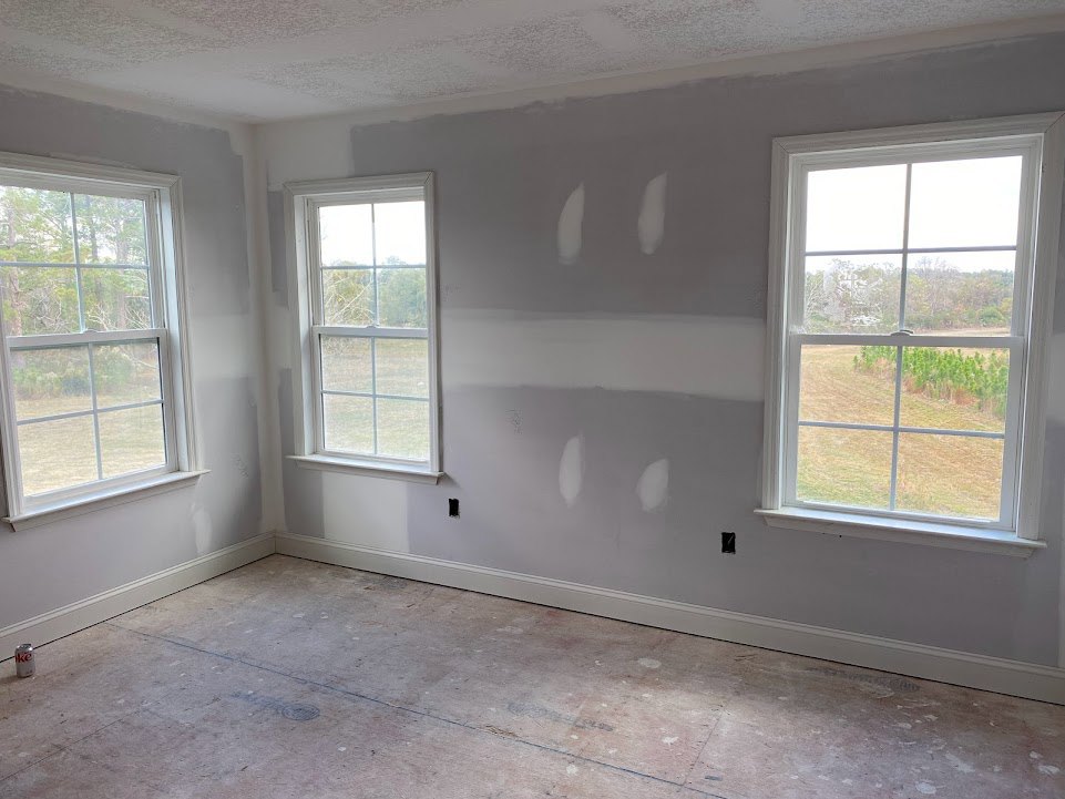 White-walled room featuring large windows overlooking trees, concrete floor with visible dust and baseboard molding