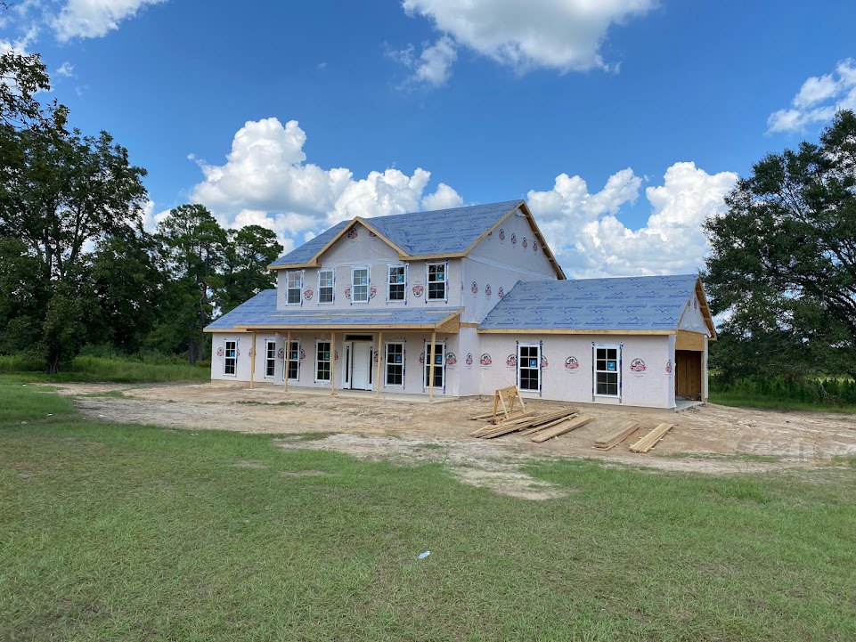 Wood-framed house under construction on grassy lot, partially completed roof, scattered wooden planks, blue sky with clouds, leafy tree nearby