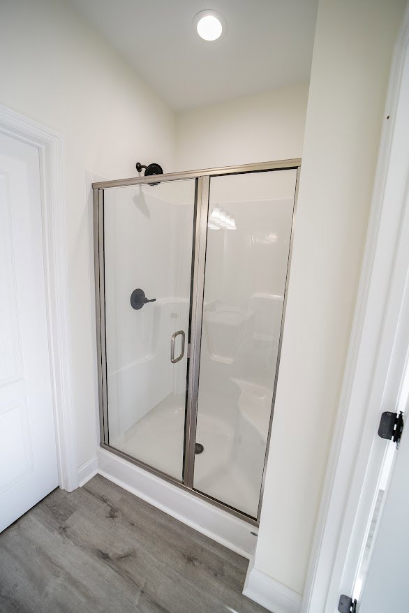 Walk-in shower with clear glass doors, light tile walls, chrome door handle, plaster ceiling, and partial bathroom mirror visible